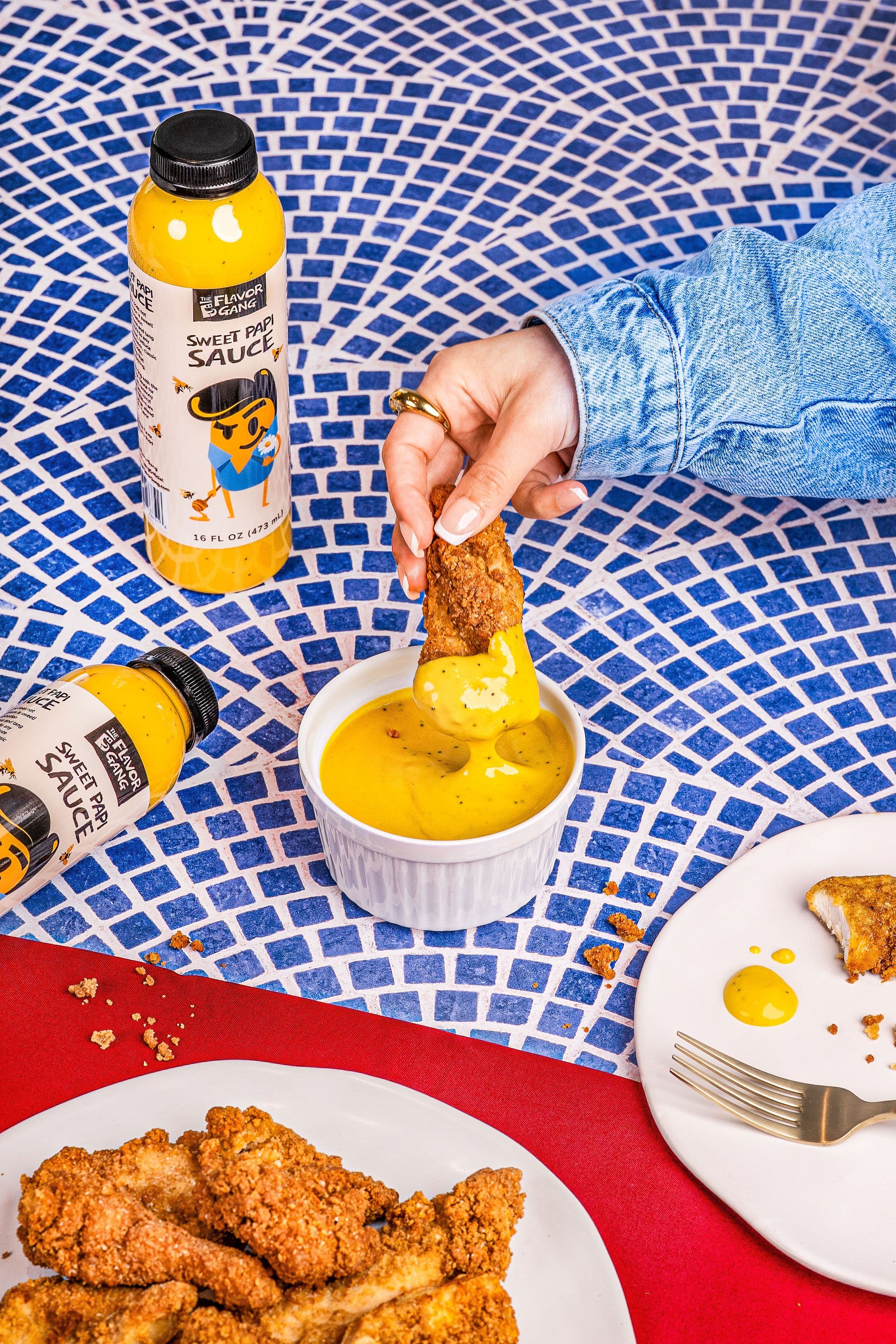 Hand dipping a crispy chicken tender into a bowl of The Flavor Gang Sweet Papi honey mustard sauce on a blue tile background.